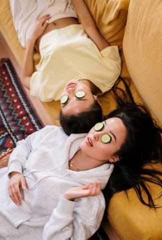 An image of two women lying down on the sofa with cucumber slices on their eyes