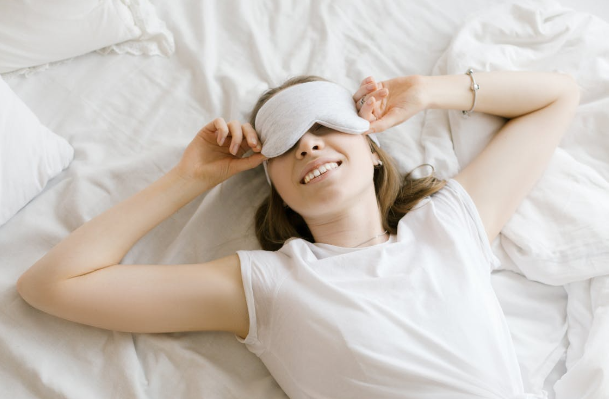 A young woman lying in bed with an eye mask, smiling and appearing relaxed.